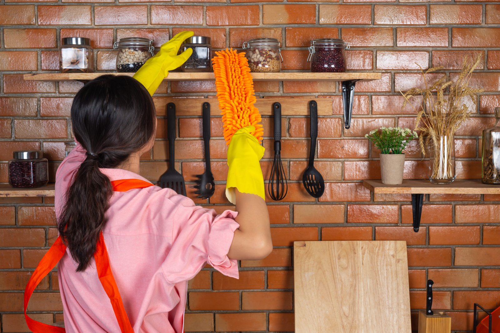 Young girl is wearing yellow gloves while cleaning the kichen room with duster in her house.